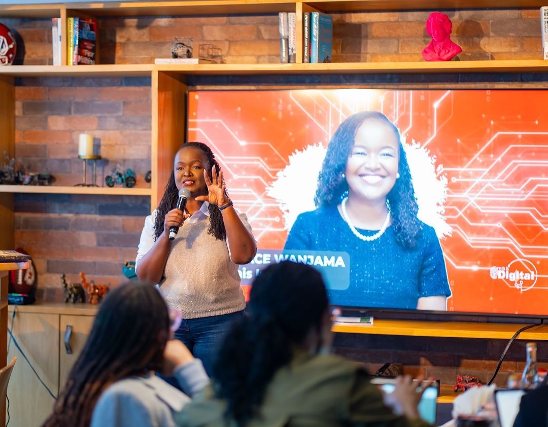 Speaker presenting on stage with a TID red screen displaying the name "Grace Wanjama" behind them
