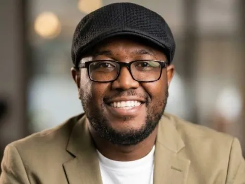 Philip Muigai smiling in a flat cap, glasses and tan blazer over a white T-shirt
