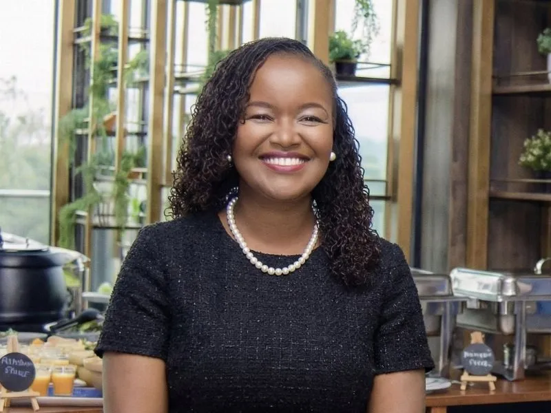 Grace Murugi smiling in a black tweed dress and pearl necklace in a bright, plant-lined venue