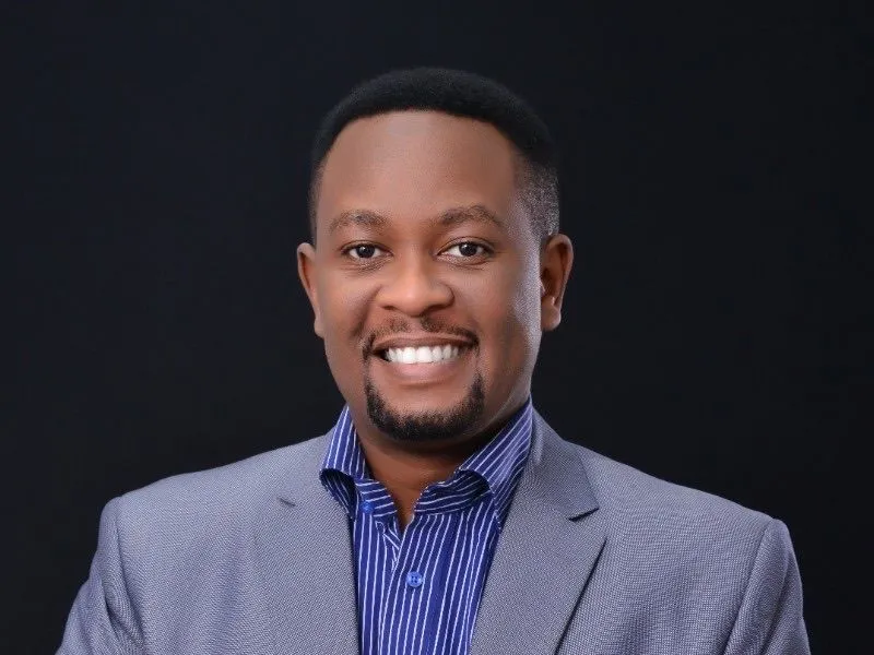 Greg Wanjama smiling in a grey blazer and blue striped shirt against a dark studio background